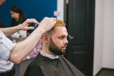 Barber Making Haircut Bearded Man In Barbershop. Professional stylist cutting client hair in salon. Barber using scissors and comb. Skillful hairdresser cutting male hair. Hair Care Service Concept.の写真素材