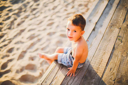 Little Caucasian boy child sitting on wooden pier sandy beach, summer time, sea vacation near water. The theme is the flow of time, a short life, the meaning and purpose of existence. Meaning of life.の写真素材