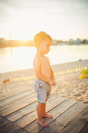 Little Caucasian boy child sitting on wooden pier sandy beach, summer time, sea vacation near water. The theme is the flow of time, a short life, the meaning and purpose of existence. Meaning of life.の写真素材