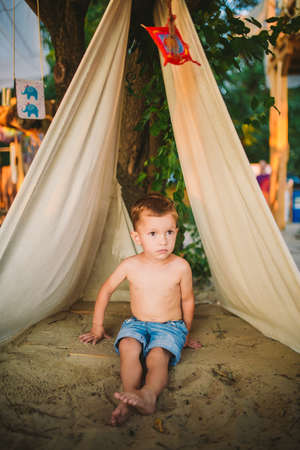 theme summer vacation, little boy, Caucasian child playing in wooded area in park on playground in the yard. The kid in Tipi wigwam tent in summer. Explore and play in nature in the summer time.の写真素材