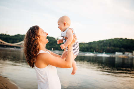 mother which turns the child against a sunset and water. Happy mom and baby. Playing on beach. Young woman tossing up her son.の写真素材