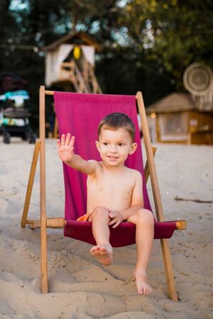 Beach boy. Little beach baby sitting on beach chair. Boy sitting on the chair by sea. Little boy sitting alone on the chair on the beach and looking at sea waves.の写真素材
