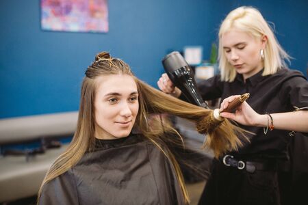 Portrait of happy woman at the hair salon. Professional hair styling concept. Hairdresser drying girl long hair using hairdryer and brush. Drying With Blow Dryer.の写真素材