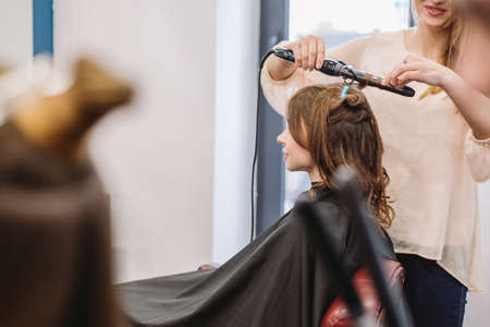 beauty, hairstyle concept, happy young woman and hairdresser with hair iron making hairdo at hair salon. Woman Having Hair Stylized Hairdresser. Smoothes Hair Curling. Stylist Using Tool for Modeling.の写真素材