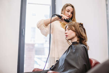 beauty, hairstyle concept, happy young woman and hairdresser with hair iron making hairdo at hair salon. Woman Having Hair Stylized Hairdresser. Smoothes Hair Curling. Stylist Using Tool for Modeling.の写真素材
