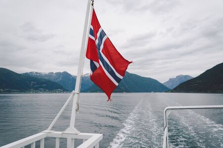 Norwegian flag hanging on the railing of the ship and waving above the water. Norvegian fjord with a flag. Ferry trip in Norway. Norway Flag on sea and mountains background.の写真素材