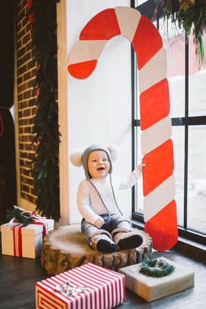 New year and Christmas holidays theme Caucasian child boy 1 year old sitting on a stump felled tree near the window in a funny hat. Scenery large red striped candy.の写真素材
