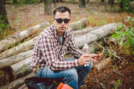 Fast food theme snack on nature. Caucasian man eating a sandwich and drinking water from a palette bottle in the forest. Tourist stopped for lunch in a wooded area.の写真素材