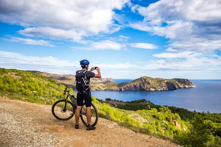 a young guy on a mountain bike trails in Spain and takes a photo on a white phone in the background of the Mediterranean sea of the rocky coast of the Costa Brava. In a blue helmet on his head and a black backpack. Sunny weather on a mountain road near the city of Cadaquesの写真素材