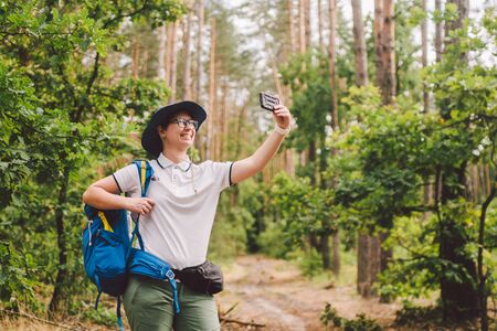 Smiling female tourist holds smart phone, takes selfie against beautiful landscape wood. Hiking woman with backpack taking selfie photo with smartphone. Travel and healthy lifestyle outdoors.の写真素材