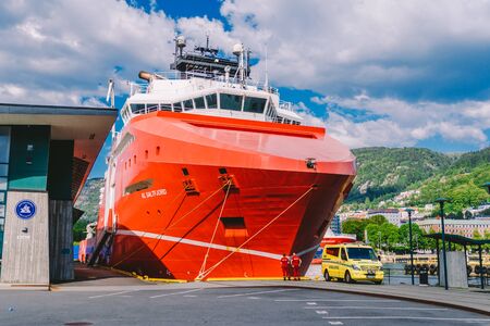 Two Norwegian paramedics in red uniforms are resting near an ambulance parked in a port near a large...