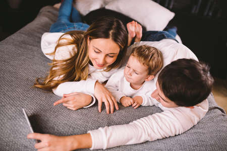 A young family lies on the bed and looks at the mobile phone. Mom, dad and son are watching a video on a smartphone in the bedroom in the evening. Family evening vacation at home.の写真素材