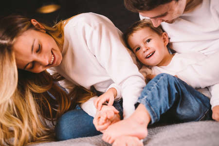 happy family is having fun in bedroom. Enjoying being together. Parents are tickling their little son while lying in bed. Cheerful family playing together on bed. Smiling Family Having Fun.の写真素材