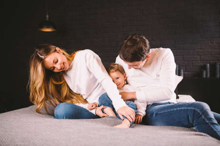 Parents with their baby boy on bed at home. Families with child in bed at night at home. Happy family enjoying in bed. Mom, dad and kid play in the bedroom in the evening. Loft style interior.の写真素材