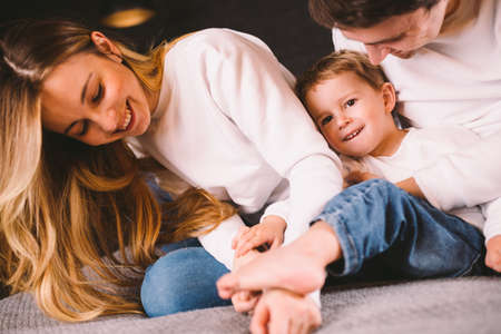 Happy family having fun lying down on bed at home. Happy Family In Bed. Mom dad and son lie on bedroom at within doors. father, mother and child relaxing bedstead in loft style apartment in evening.の写真素材