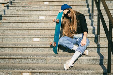 Young caucasian woman posing on street with skateboard in hands. Teenager girl in blue jeans extreme sports in an urban environment. Theme of youth recreation, lifestyle. Skateboarder in the city.の写真素材