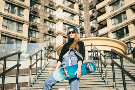 Urban woman with skate. Hipster girl with skateboard in city. Extreme sport and emotions concept. Alternative lifestyle. Stylish hipster girl holding skateboard and posing. Street style outfit.の写真素材