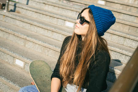 Stylishly dressed woman in blue denim jumpsuit posing with skateboard. Street photo. Portrait of...