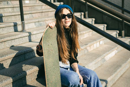 Young caucasian woman posing on street with skateboard in hands. Teenager girl in blue jeans extreme sports in an urban environment. Theme of youth recreation, lifestyle. Skateboarder in the city.の写真素材