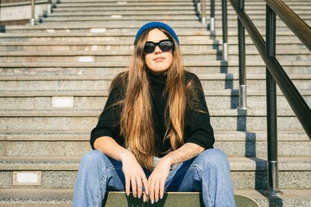 Stylishly dressed woman in blue denim jumpsuit posing with skateboard. Street photo. Portrait of girl holding skateboard. Lifestyle, youth concept. Leisure, hobby and skate in the city.の写真素材