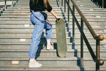 Urban woman with skate. Hipster girl with skateboard in city. Extreme sport and emotions concept. Alternative lifestyle. Stylish hipster girl holding skateboard and posing. Street style outfit.の写真素材