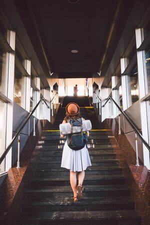 Young beautiful caucasian woman tourist in a summer dress and hat with a tourist backpack climbs up the summer woman at the railway station. Student girl travels. Theme trip and train station.の写真素材