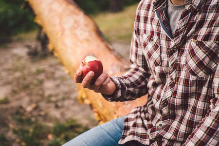 The theme is healthy eating and vegetarianism. Outdoor activities in the forest and fruit snack. Young Caucasian male hiker resting and eating an apple in a clearing near a forest lake.の写真素材