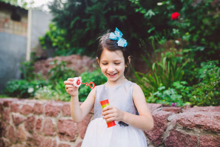 Little Caucasian girl in a white dress plays with soap bubbles in the backyard of the house on the green grass. Childhood and joy to small things. Summer and weekend.の写真素材