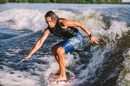 Young athletic man with long hair wakesurfing on waves of river in sunny summer weather. Ttheme outdoor activities in summer. Water sports wakesurf on the board. sliding wakeboarder in water splash.の写真素材