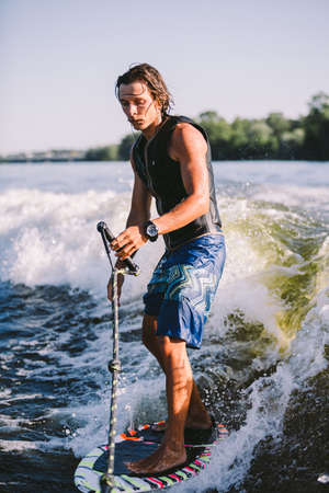 Young athletic man with long hair wakesurfing on waves of river in sunny summer weather. Ttheme...
