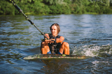 Young athletic man with long hair wakesurfing on waves of river in sunny summer weather. Ttheme...