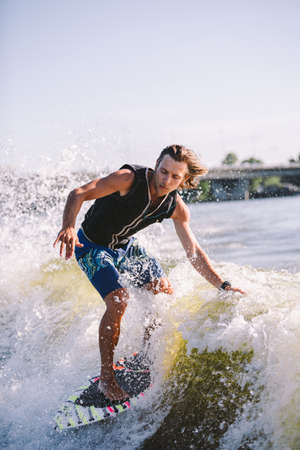 Beautiful long haired blond wakesurf in vest on board along waves of lake. Athletic male athlete wakes surfing in summer on river. Wakesurf water sport. Surfer on wave. Man surfing on the Surfboard.の写真素材