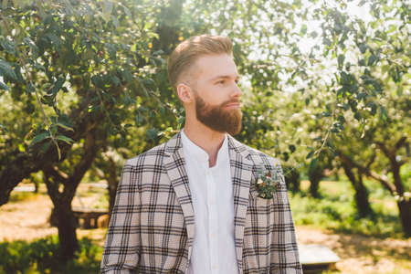 Portrait of Caucasian stylish bearded groom in vintage suit posing outdoors in sunny weather. Handsome man with beard in plaid jacket. Male model at wedding in rustic style. Groom in checkered jacket.の写真素材