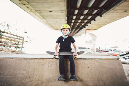 Boy in helmet posing with skateboard. Portrait stylish lad child with skate board outdoors. Theme sport, children health. Caucasian child skateboarder posing with skate board in outdoor skate park.の写真素材