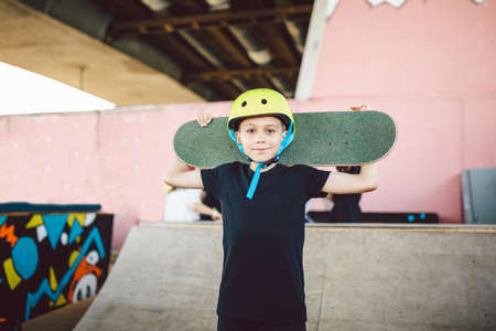 Caucasian child skateboarder posing with skate board on shoulders an outdoor skate park. Portrait of boy is engaged in skateboarding on half pipe ramp. Outdoor activities and extreme sports in summer.の写真素材
