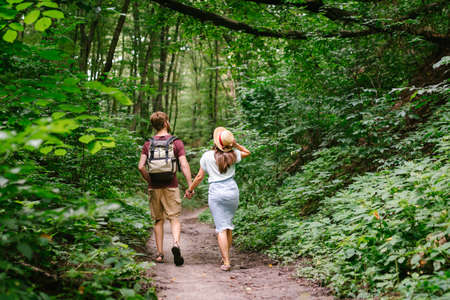 Happy caucasian couple are walking in a dense forest along the path holding hands, rear view. Hikers with backpack looking for place for picnic in wooded area in the summer. Walk outdoors together.の写真素材