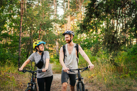 Young caucasian couple of cyclists walk and push their mountain bikes along the forest road in the...