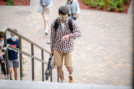 Man Carrying folded E-Scooter in hand on stairs near building. Moving by eco urban transport, modern city concept. Ecological technological lifestyle. Person Carry Electric Scooter In Folded Position.の写真素材