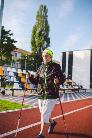 Active rest of the elderly theme. Sports and health in retirement. Caucasian very old woman with deep wrinkles doing Nordic walking exercises with sticks outside in the city stadium.の写真素材