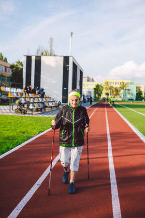 Active rest of the elderly theme. Sports and health in retirement. Caucasian very old woman with deep wrinkles doing Nordic walking exercises with sticks outside in the city stadium.の写真素材