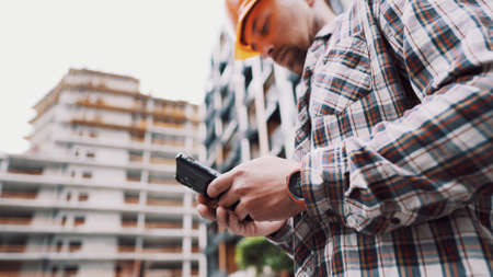 Inclusion of mobile applications in the development management process. Caucasian engineer builder in hard hat use on construction site mobile phone, engineer at work checking building project.の写真素材