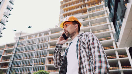 Caucasian male construction worker in orange hard hat and plaid shirt talking on phone at construction site. Architecture theme. Male profession. Foreman controls construction process by smartphone.の写真素材
