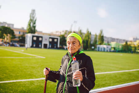 Old woman walking with nordic walk sticks on running track, rubber treadmill and stopped to quench thirst, drink water from a flask. Active female senior. Active retirement, healthy lifestyle concept.の写真素材