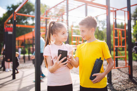 Caucasian children teenagers use cell phone in workout at street gym. Diverse twins watching video...