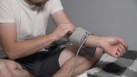 Young caucasian man measures blood pressure at home in bedroom sitting on bed at home clothes. Male uses medical instrument tonometer to check his own blood pressure. Hypertension at young age.の写真素材