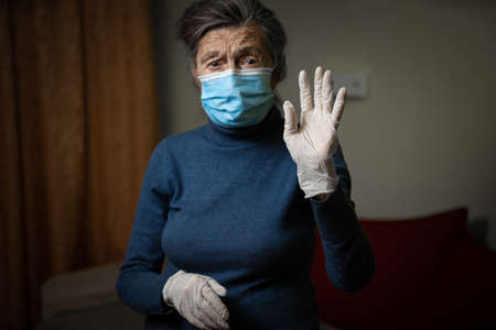 White medical glove in foreground, shown by an elderly grandmother wearing a medical mask, calling for safety and sanitation during an epidemic. Senior woman in personal protective equipment.の写真素材