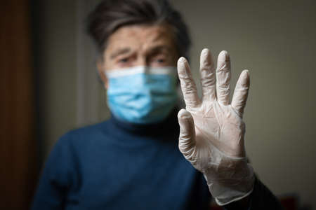 Importance wearing gloves and masks to protect of coronavirus, especially elderly and seniors. Focus on latex gloves on background old woman in medical mask. Sign indicates protection against covid 19の写真素材