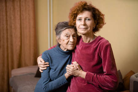 Adult daughter takes care of elderly mother suffering from dementia, old woman ninety years age gray hair and wrinkles on face and sweet kind smile, family idyll caring for elderly, hug each other.の写真素材