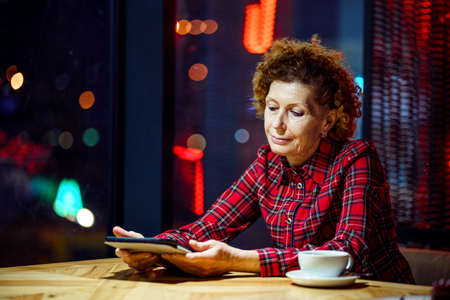 Mature woman with blond curly hair checks mail and social media notifications via tablet, smiling female holding tablet, watching video, surfing internet inside coffee shop in evening by window.の写真素材
