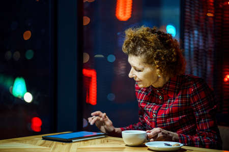 Mature caucasian woman drinking coffee and watching an electronic tablet, use internet. sitting...